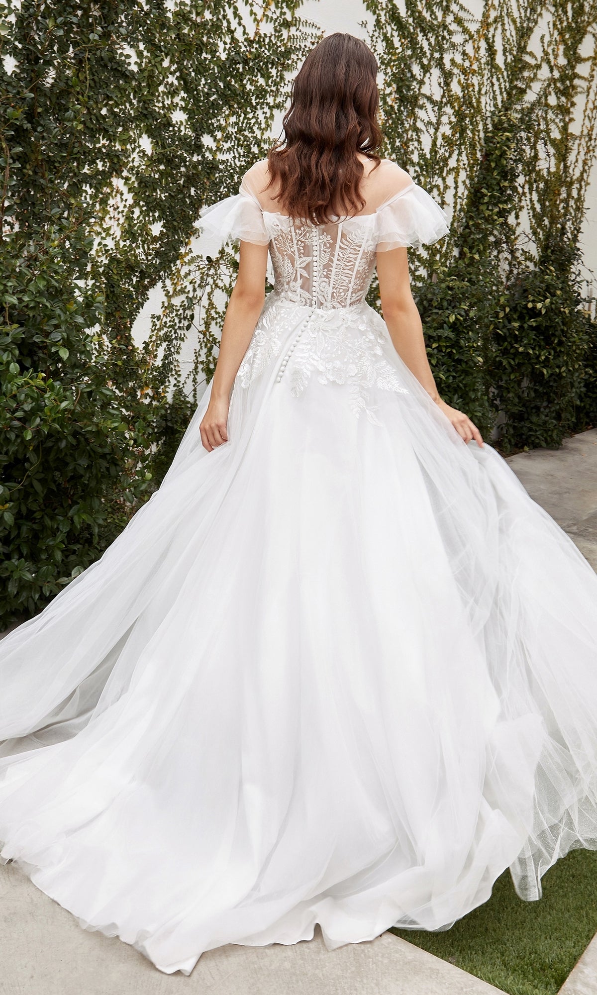 A woman stands outdoors surrounded by greenery, wearing the Ladivine White A-Line Wedding Dress A1070W featuring ruffle off-the-shoulder sleeves and a lace-up back.